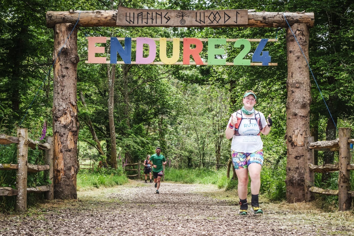 Photo of Ruth running through a wooden gateway in the forest which has the words 'wasing woods' etched in norse-like words above, and a colourful 'ENDURE24' sign. Ruth is smiling at the camera with both thumbs up, wearing a white tshirt which has her name on it, and 'Fundraising for Breathworks'.