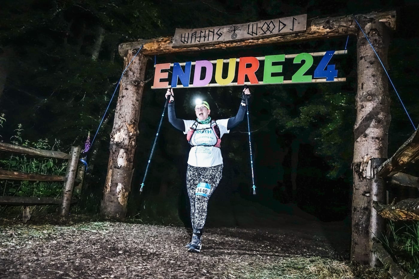 Photo of Ruth at night in the dark coming through the Wasing Woods gate. She's got a headtorch on her head which is switched on, and reflective leggings which are showing bright white dots. She's holding both her poles up in the air in an expression of delight.