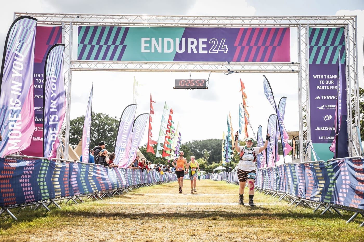 Photo of Ruth coming over the finish line for the last time, the clock shows the time of 10:26:52 and she's holding both hands up in triumph and smiling at the camera.