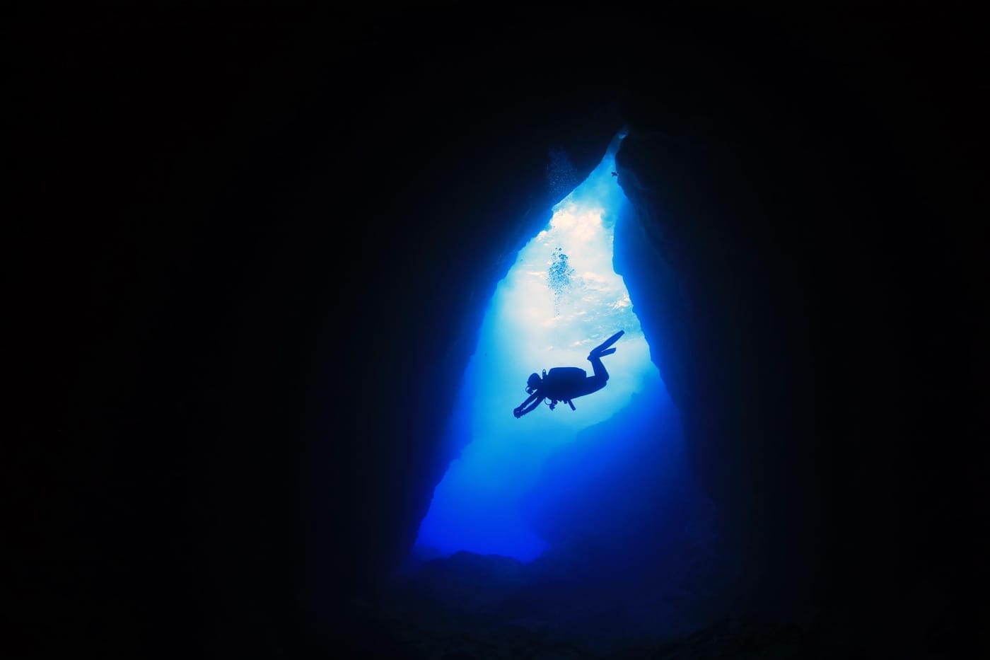 A photo of a scuba diver descending into a dark cave, looking up to the surface.