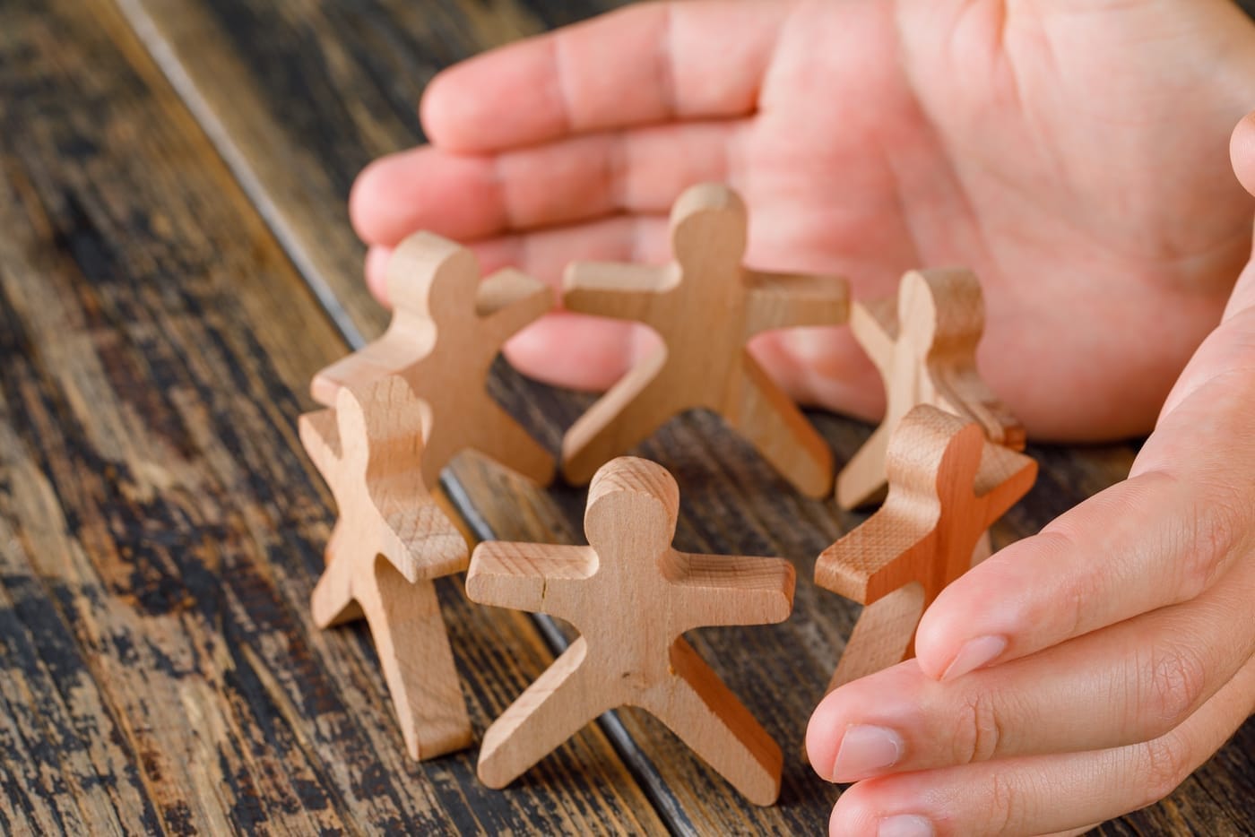 A wooden table with six wooden figures of people in a circle appearing to hold hands. There's a person with two hands cupping around one side of the circle of figures in a supportive looking stance.