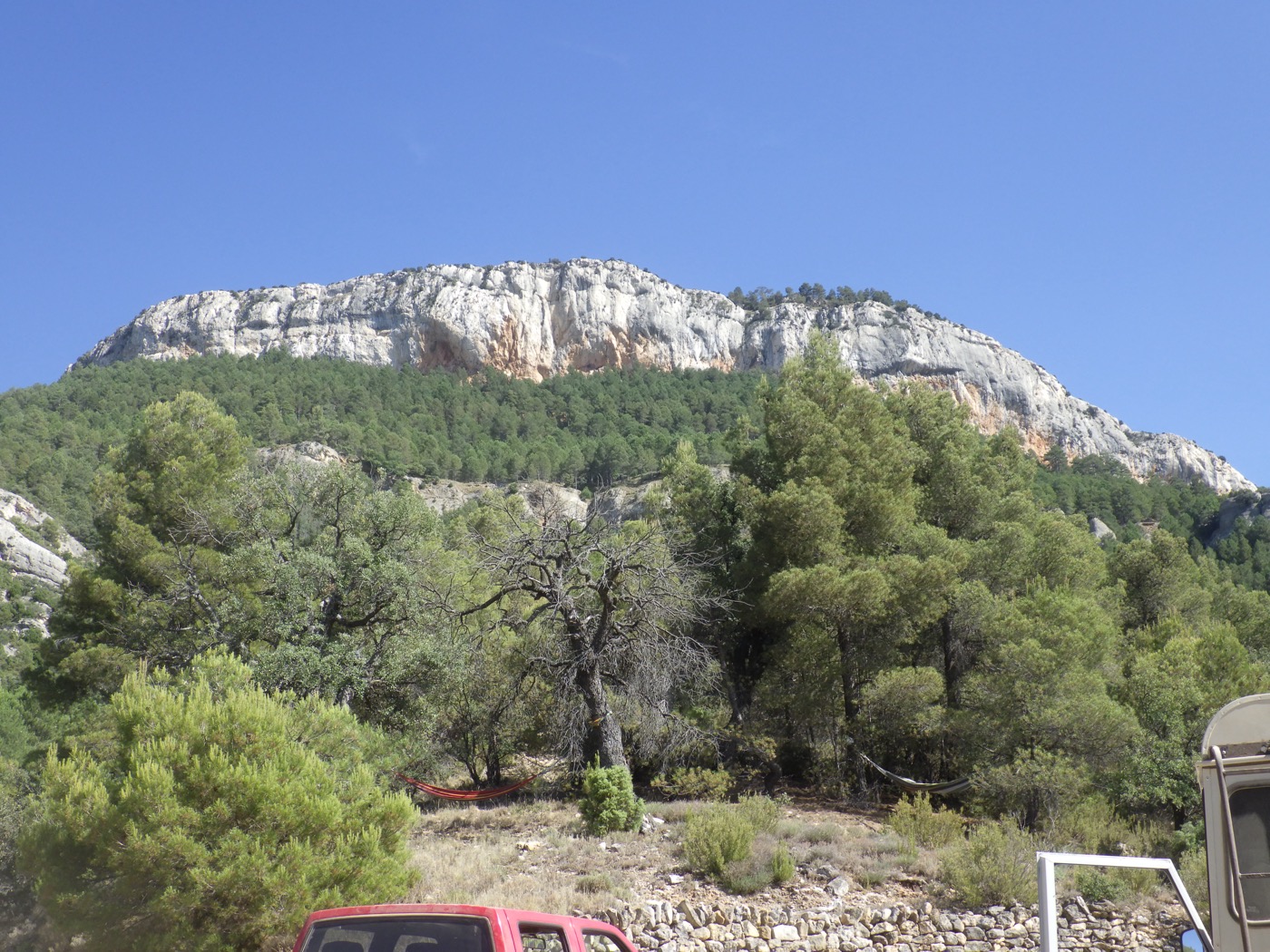 The white limestone cliff of the Akashavana massif rising above pine forest under a clear blue sky, with a hammock strung between trees in the foreground.