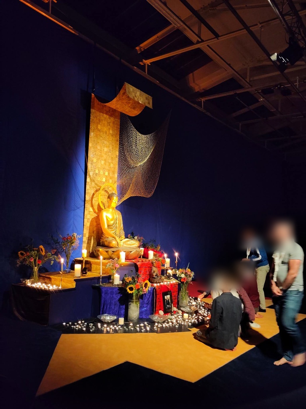 A large golden Buddha rupa on a stage, surrounded by flowers and hundreds of tea lights, lit against a deep blue backdrop.