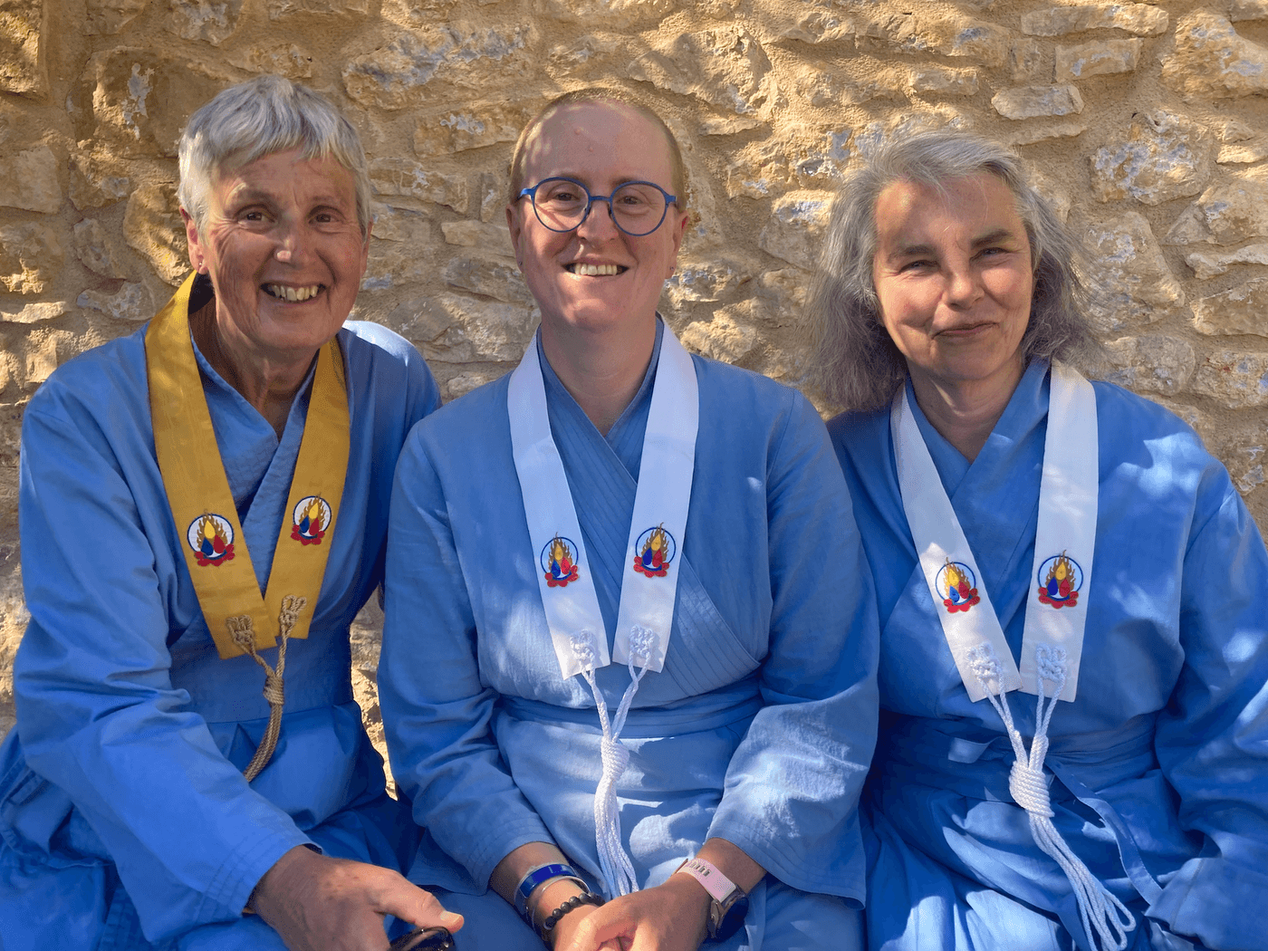 A photo of three women wearing blue robes, Sīlavāpi in the middle with a white strip of cloth called a kesa around her neck with the symbol of the Triratna movement - a yellow, red and blue drop on a lotus surrounded by a ring of flames - the woman on the left is wearing a saffron coloured kesa. Both women on the left and right of Sīlavāpi are much older. All three are smiling to the camera.