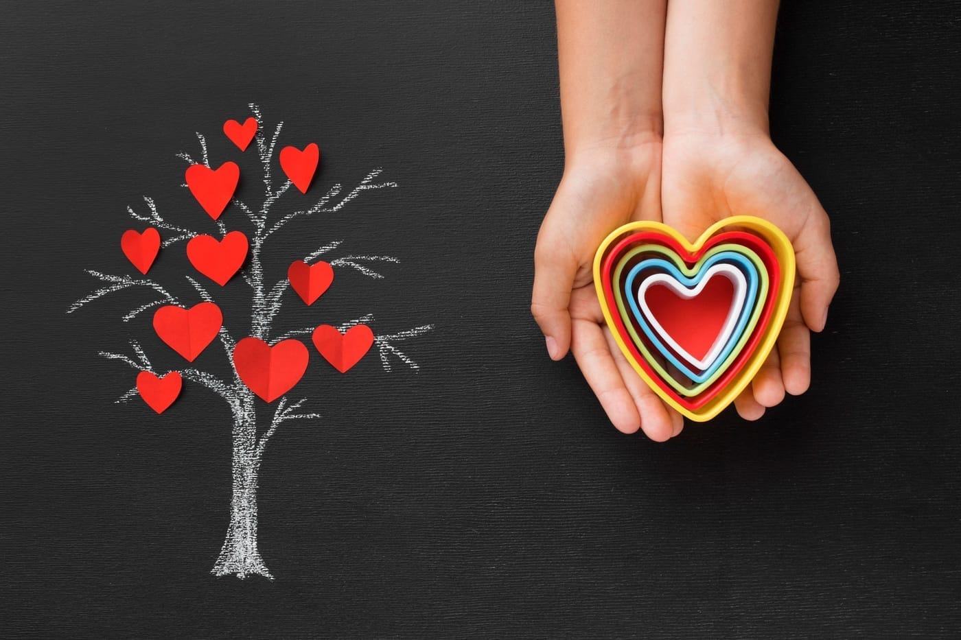 A pair of hands holding a colorful layered heart sits on the right side of the image. On the left, a chalk drawing depicts a tree with red hearts as leaves. The background is dark, enhancing the contrast between the elements.