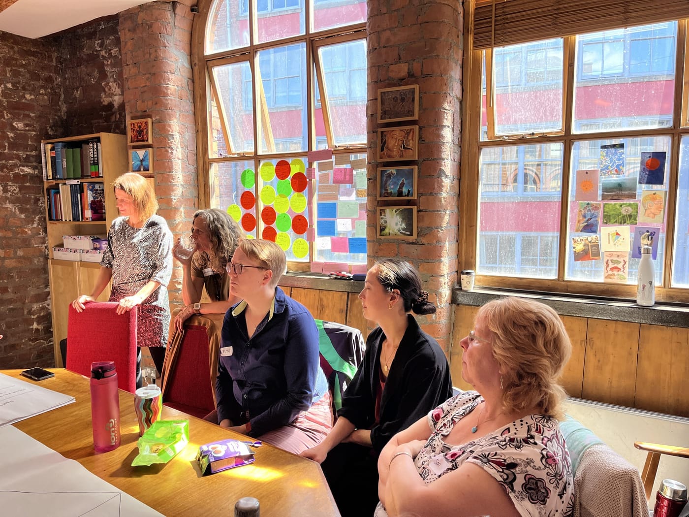 Members of the Breathworks board and team seated around the table, listening, beside tall brick-framed windows covered in notes and postcards.