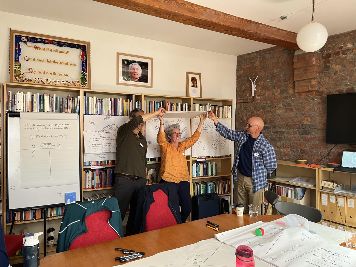 Three of the group standing with their arms raised and hands joined above their heads, laughing, in front of the Breathworks library shelves.