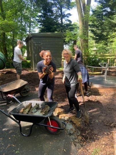 Two volunteers grinning at the camera beside a wheelbarrow of soil, one holding a clump of earth, while others dig behind.