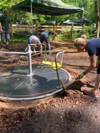 Volunteers digging out the ground around an accessible playground roundabout, one kneeling in gardening gloves, under a gazebo in the woods.