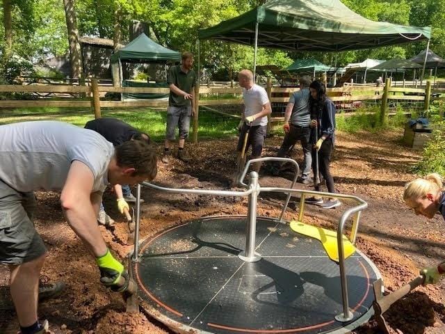 A team of volunteers digging around the playground roundabout, with green gazebos and woodland behind.