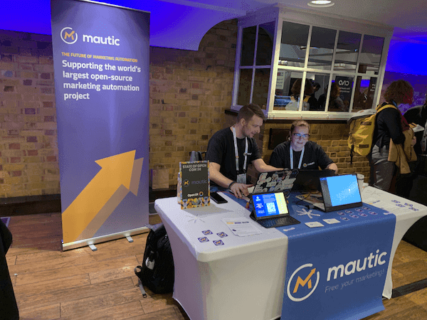 The Mautic stand at State of Open Con, with a rollup banner to the rear left, a white covered table with a blue roller which has the Mautic logo on it, and a screen showing a video of Mautic. The tablet also shows a landing page with a QR code. Two people are sitting behind the stand.