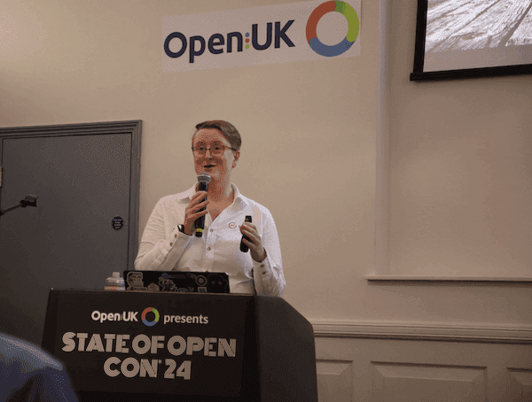 Photo of Ruth wearing a white, long sleeved shirt with the Mautic logo embroidered on it, holding a microphone and speaking at Open UK.